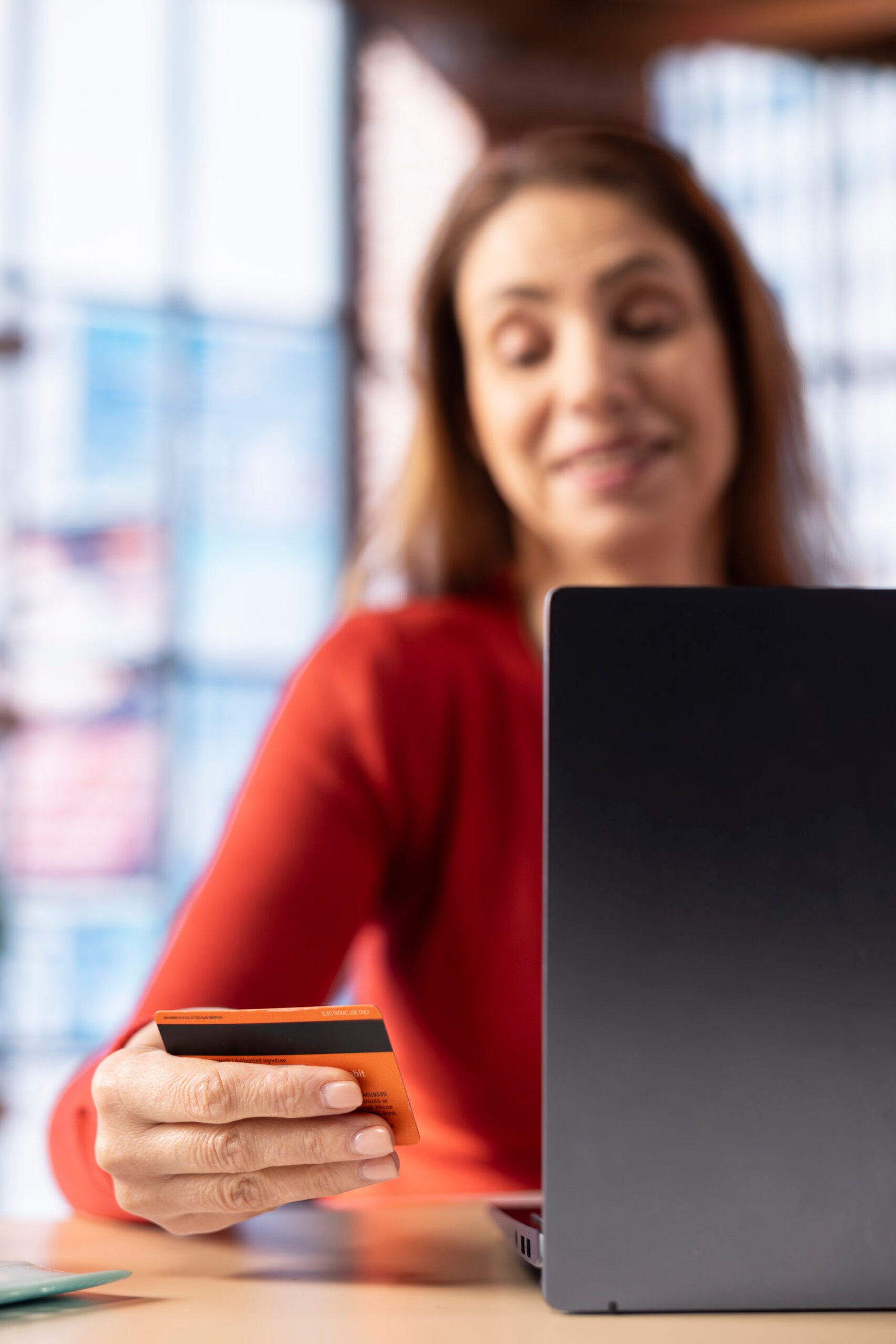 Adult holds her card to check balance and oversee budget planning, uses laptop at home office to track her expenses responsibly and confirm payments. Mature woman manages her finances.